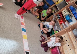 Dzieci skaczą obok obrazków rozłożonych na dywanie i nazywają właściwy kolor po angielsku.