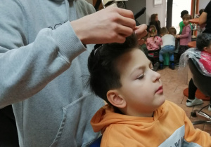 Szymin siedzi na fotelu fryzjerskim, ma robioną fryzurę
