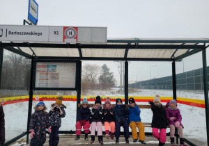 Dzieci stoją i siedzą na przystanku, czekają na autobus