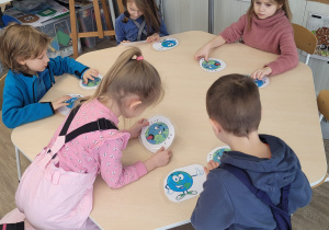 Dzieci siedzą przy stolikach, mają w ręku karty potrzebne im do zadania