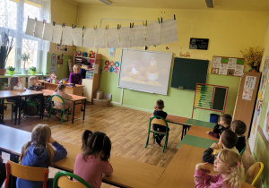 Dzieci siedzą w ławce szkolnej i oglądają bajkę o wielkanocy na tablicy interaktywnej