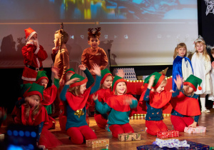 Elfy siedzą na scenie wykonują układ taneczny z pudełkami