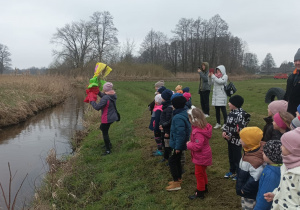 Dzieci wraz z Paniami stoją nad brzegiem rzeczki. Pani Kasia wrzuca Marzannę do wody