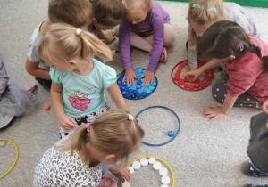Dzieci siedzą na dywanie, układają kolorowe korki według koloru kółek ringo.