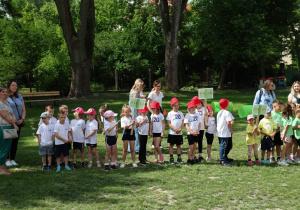 Dzieci w czerwonych czapeczkach i białych koszulkach z logo przedszkola stoją do ogłoszenia startu olimpiady