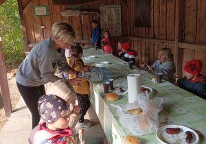Dzieci przy stole jedzą kiełbaski z ogniska.
