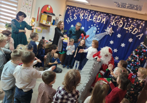 Dzieci stoją, śpiewają piosenkę dla Mikołaja, a Św. Mikołaj siedzi na czerwonym tronie przy choince, wyjmuje prezenty z worka