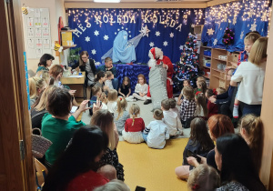 Dzieci siedzą na dywanie, rodzice ogladają rozdawanie prezentów przez św.Mikołaja. Mikołaj zasiadł przy choince.
