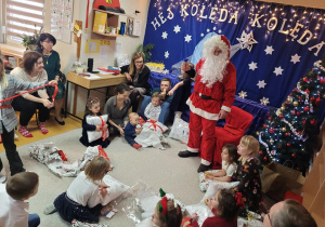Dzieci z prezentami siedzą na dywanie, Mikołaj stoi