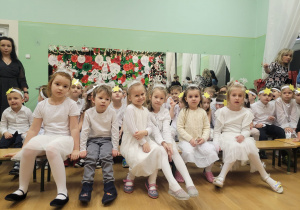 Dzieci ubrane na biało siedzą na ławce podczas przerwy między występami