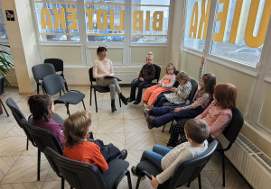 Dzieci siedzą na krzesełkach w półkolu, słuchają wiersza